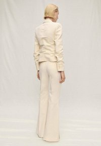 Beige přizpůsobený blazer se strukturovanými rameny, spárovaný s kalhotami s vysokým pasem a rozšířenými nohavicemi, obojí v hladké látce s minimálním množstvím švů.