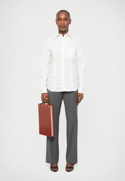 Chemise blanche à manches longues avec texture froncée, pantalon gris à jambes larges, et un dossier en cuir marron avec des accents dorés, porté avec des sandales noires.