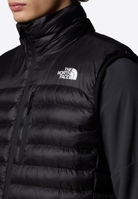 Zwarte puffer vest met hoge kraag, horizontale stiksels en een ritszak. Heeft een wit logo op de linkerborst en een gladde buitenkant.
