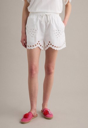 Frau trägt weiße Eyelet-Spitzen-Shorts mit gezacktem Saum und rosa Slipper mit roten und rosa Quasten, steht vor neutralem Hintergrund.