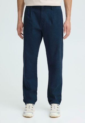 Homme portant un pantalon droit bleu foncé et des baskets blanches, debout devant un fond clair uni.