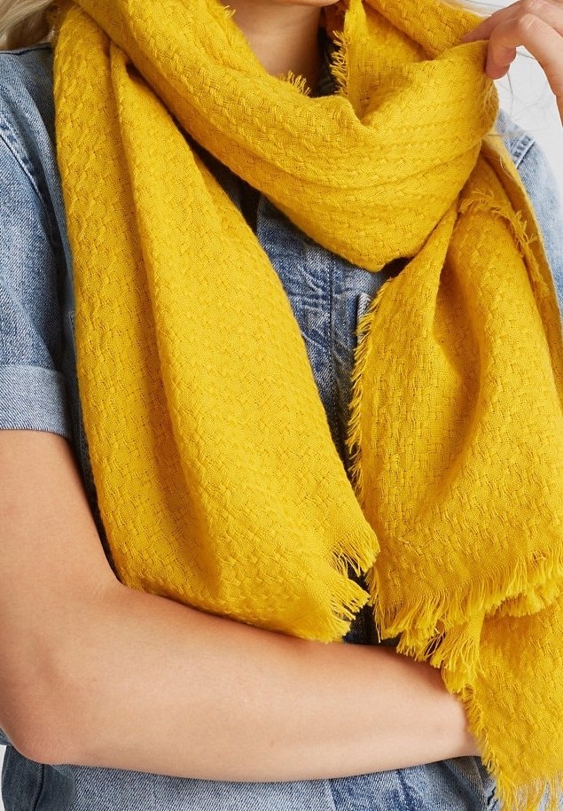 Une personne porte une grande écharpe jaune texturée par-dessus une chemise en jean à manches courtes, tenant une extrémité de l'écharpe avec la main.