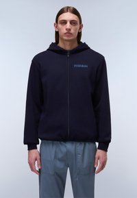 Haut de survêtement zippé bleu marine avec capuche à cordon de serrage, arborant un logo bleu sur la poitrine et des poignets côtelés, associé à un pantalon à cordon de serrage gris clair.