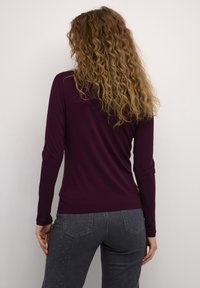 Maglietta a maniche lunghe di colore viola realizzata in tessuto morbido, con un design aderente. La vista posteriore mostra una texture liscia e un collo rotondo. Abbinata a jeans grigi.