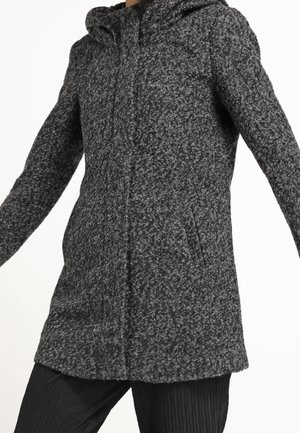 Długi ciemnoszary płaszcz z teksturowaną tkaniną i kapturem, noszony przez osobę z lekko uniesionymi ramionami, w połączeniu z czarnymi plisowanymi spodniami.