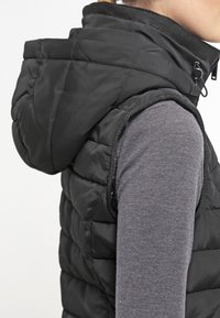 Černá péřová vesta s kapucí, s prošívaným designem a zipovým zapínáním. Nosená přes šedé tričko s dlouhým rukávem, ukazující oblast rukávu.