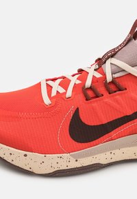 Scarpa da atletica rossa con tomaia in mesh testurizzato, accenti marroni a contrasto, lacci color crema e suola beige screziata, con logo swoosh nero.