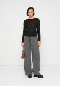 Μαύρο μακρυμάνικο τοπ με εφαρμοστή σχεδίαση, συνδυασμένο με φαρδιά γκρι παντελόνια και μαύρες μπότες με χοντρή σόλα. Κρατά ένα καφέ τσάντα.