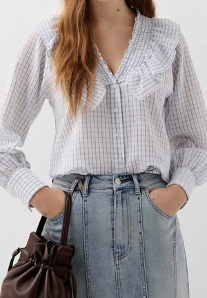Femme portant un chemisier blanc et bleu à carreaux avec des volants, rentré dans un jean en denim bleu clair taille haute, tenant un sac à main en cuir marron.