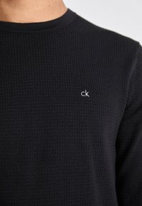 Maglione nero a maglia con un motivo testurizzato, caratterizzato da un collo rotondo e un piccolo logo bianco "ck" sul petto.