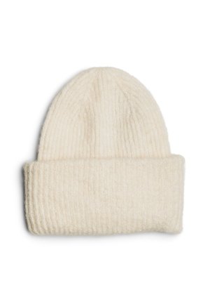 Une femme porte un manteau marron foncé à col montant et un bonnet en maille couleur crème devant un fond uni clair.