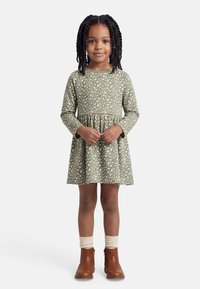 Νεαρή κοπέλα με πλεξούδες, ντυμένη με μακριά φορεματάκι χακί με σχέδια καρδιές, κρεμ κάλτσες και καφέ μποτάκια μέχρι τον αστράγαλο, στέκεται ήρεμη.