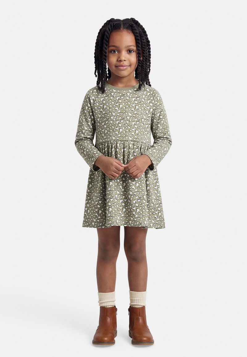 Νεαρή κοπέλα με πλεξούδες, ντυμένη με μακριά φορεματάκι χακί με σχέδια καρδιές, κρεμ κάλτσες και καφέ μποτάκια μέχρι τον αστράγαλο, στέκεται ήρεμη.