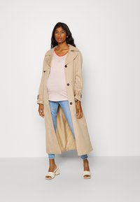 Trench coat bege com colarinho oversized, grandes botões e bolsos, usado sobre uma camisa riscada em rosa, combinado com jeans azul claro e sandálias creme.
