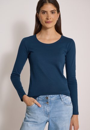 Femme aux longs cheveux bruns porte un t-shirt bleu marine cintré à manches longues et un jean taille haute bleu clair, une main dans la poche, fond neutre.