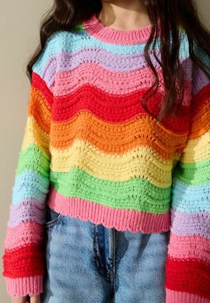 Personne portant un pull en maille rayé multicolore avec un motif ondulé et un jean en denim bleu, visible du milieu de la poitrine jusqu'aux hanches.