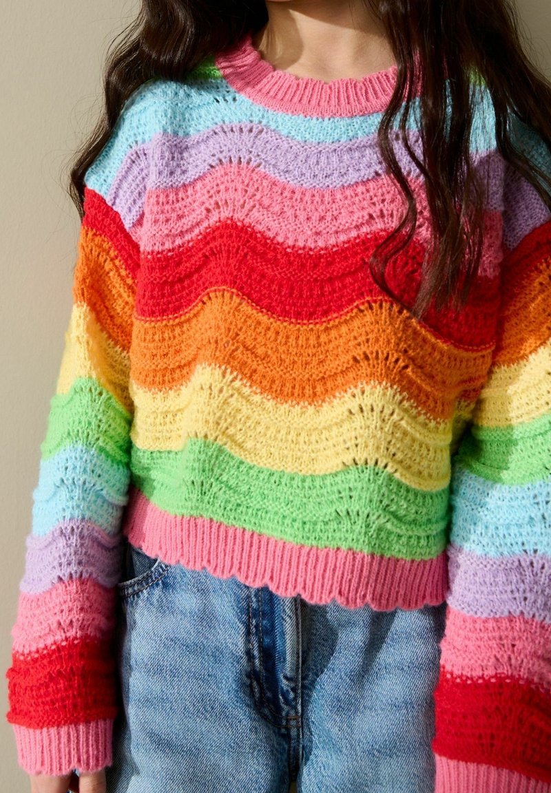 Personne portant un pull en maille rayé multicolore avec un motif ondulé et un jean en denim bleu, visible du milieu de la poitrine jusqu'aux hanches.