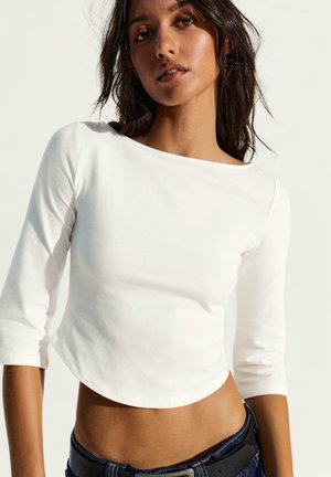 Femme aux cheveux foncés portant un haut blanc court à manches longues et un jean bleu foncé, debout devant un fond clair.