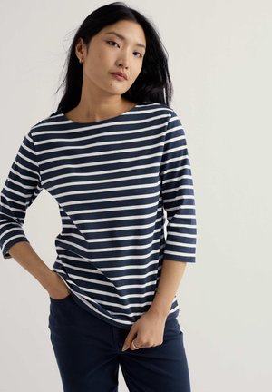 Vrouw met lang zwart haar, gekleed in een marineblauw-wit gestreept shirt met driekwart mouwen en marineblauwe broek, staand tegen een effen achtergrond.