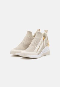 Sneakers beige in maglia con tomaia testurizzata, accenti dorati e suola bianca. Presentano una zip e un taglio sul retro per indossarli facilmente.