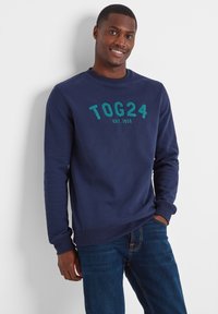 Navy blue sweatshirt with embroidered "TOG24 EST. 1958" in teal. Ribbed cuffs and hem, soft fabric, relaxed fit, paired with dark denim jeans.