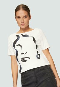 T-shirt en coton blanc à manches courtes, présentant un design de visage abstrait noir. Porté avec un jean noir décoré d'accents à clous.