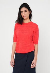 Κόκκινο πλεκτό πουλόβερ με υφασμένο διακοσμητικό μοτίβο ρόμβου, λαιμόκοψη με κυματιστό σχέδιο και κοντά μανίκια, σε συνδυασμό με σκουρό μπλε φούστα.