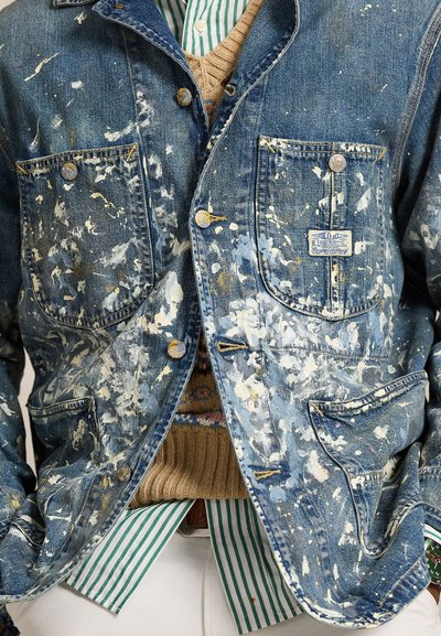 Τζιν μπουφάν με λευκές πιτσιλιές μπογιάς πάνω από ριγέ πράσινο και λευκό πουκάμισο, κάστανο πλεκτό γιλέκο και λευκό παντελόνι.