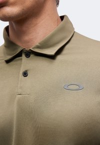 Homme portant un polo vert olive avec deux boutons noirs et un petit logo ovale gris sur la poitrine sur un fond uni.