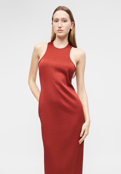 Γυναίκα με μακριά ίσια μαλλιά, που φοράει αμάνικο, εφαρμοστό κόκκινο φόρεμα, στέκεται μπροστά σε ένα απλό φωτεινό φόντο.