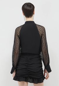 Schwarze Bluse mit durchscheinenden Spitzen-Ärmeln und einem taillierten Schnitt. Der Rücken zeigt eine glatte Textur mit einem gerafften Bund und einem Rüschensaum.
