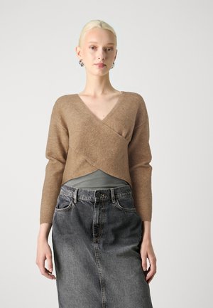 Maglione marrone avvolgente con maniche lunghe, scollo a V, texture morbida e un look a più strati sopra una maglietta grigia. Abbinato a una gonna in denim grigia.