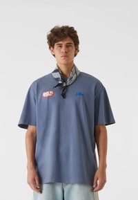 Blå bomull T-skjorte med korte ermer og løs passform, med røde "Amalfi" og blå "Saba" tekstlogoer. Brukt med et grått mønstret skjerf.