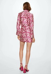 Mujer vestida con un vestido blazer de manga larga con patrón rosa y zapatos de tacón cuadrado rosa, de pie con la espalda frente a la cámara.