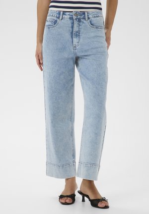 Femme portant un jean large bleu clair avec des ourlets retroussés et des sandales à talons noires, debout devant un fond blanc.