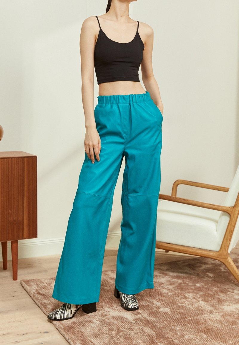 Femme portant un pantalon large turquoise, un débardeur court noir et des bottines à motif noir et blanc, debout à l'intérieur près d'une chaise et d'un meuble.