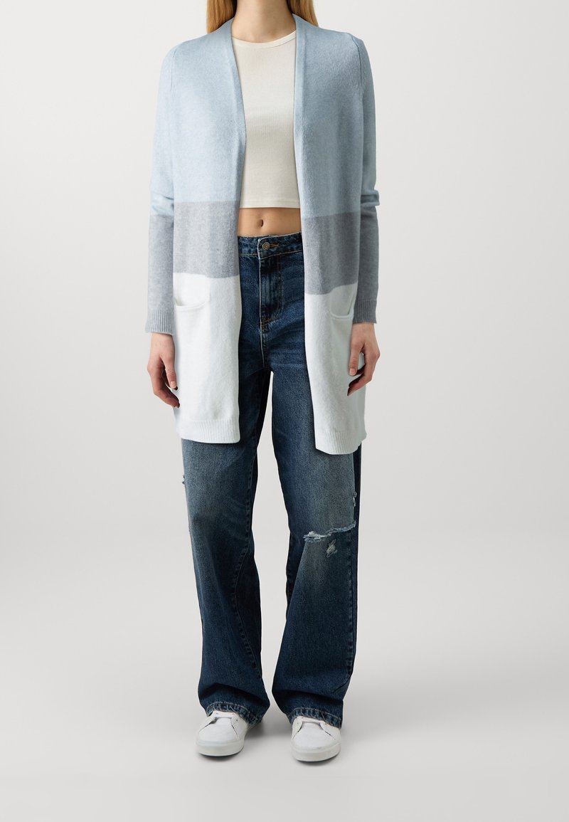 Světle modrý pletený cardigan s šedými a bílými pruhy, který má dvě přední kapsy, nošený přes bílý crop top a roztrhané džíny.