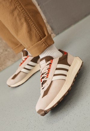 Person iført beige og brune Adidas-sneakers med hvide striber, orange snørebånd, hvide sokker og opkrogede brune bukser på en priklet gulv.