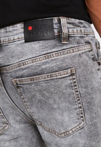 Nahaufnahme einer hellgrauen Denim-Jeanshintertasche mit brauner Naht und schwarzem Lederetikett mit der Aufschrift "DENIM PROJECT" sowie einem roten Punkt.