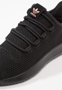 Svart idrottssko med en texturerad mesh-ovandel, svarta elastiska snören och en rosa Adidas-logotyp på tungan. Har en tjock svart sula.