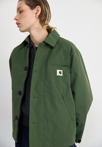 Veste imperméable verte avec un design à col, fermeture à boutons sur le devant, poches latérales et un petit logo brodé. Couche intérieure texturée visible.