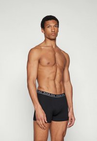 Modelo masculino con cuerpo atlético que lleva puesto un calzón negro con la cintura con el logo "PIER ONE", de pie contra un fondo claro y liso.