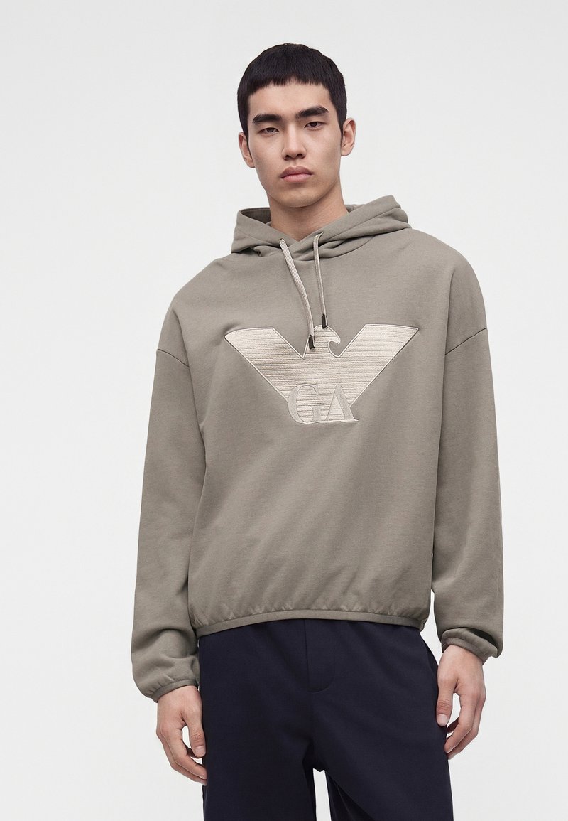Jeune homme portant un sweat à capuche taupe avec un grand logo aigle en relief et un pantalon bleu marine, debout devant un fond blanc uni.