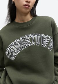 Osoba v olivovo zelenom pulóvri s veľkým bielym nápisom „GEORGETOWN“ na hrudi, so viditeľnými perami a čiernymi vlasmi.