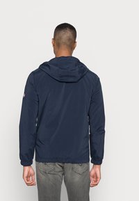 Navy windbreaker jacket with a hood, elastic cuffs, and waistband. Smooth texture, durable material, and logo detail on the sleeve.