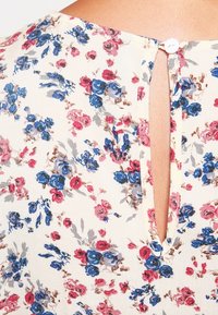 Blusa floral con una mezcla de patrones en rosa y azul sobre un fondo crema, con un escote en forma de lágrima y cierre de botón en la parte posterior.