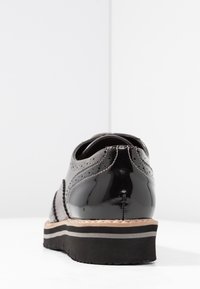 Chaussure en cuir verni noir avec un bout rond et un design à motif papillon. Dispose d'une semelle noire texturée avec une bande d'accent beige.