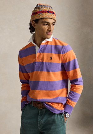Jeune homme aux cheveux bouclés portant un maillot de rugby à manches longues rayé orange et violet, un bonnet tricoté multicolore, un pantalon vert et une montre-bracelet, mains dans les poches.
