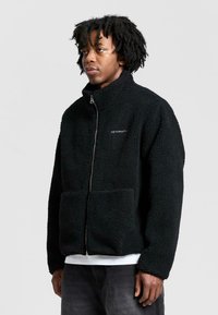 Mustat fleece-takit, joissa on korkea kaulus, vetoketjuedessä ja kaksi etutaskua. Pintakuviointi on teksturoitu. Brändin nimi "RETERNITY" valkoisella.