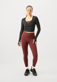 Zwarte lange mouw crop top met een diepe V-hals, gecombineerd met hoogopgetrokken bordeauxrode leggings. Model draagt zwarte sneakers met witte sokken.